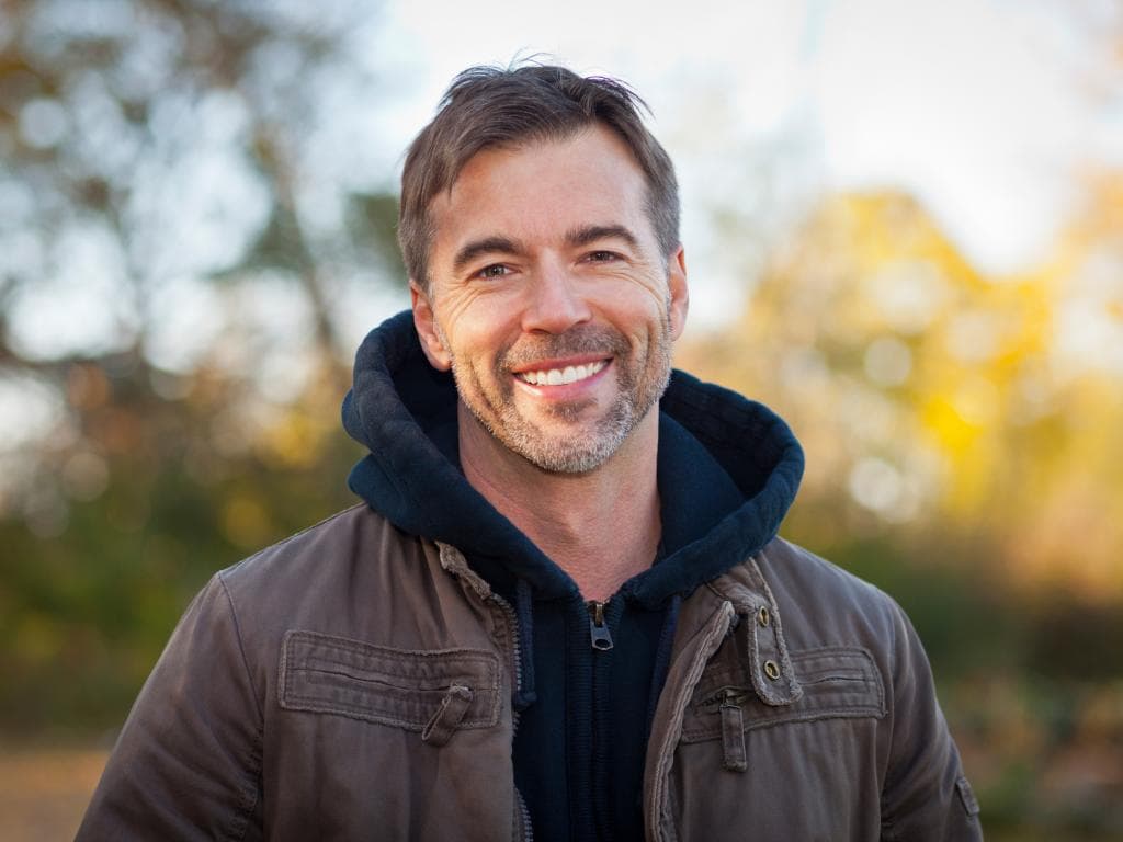 En mann i hettegenser smiler mens han står i en park, omgitt av grøntområder og trær