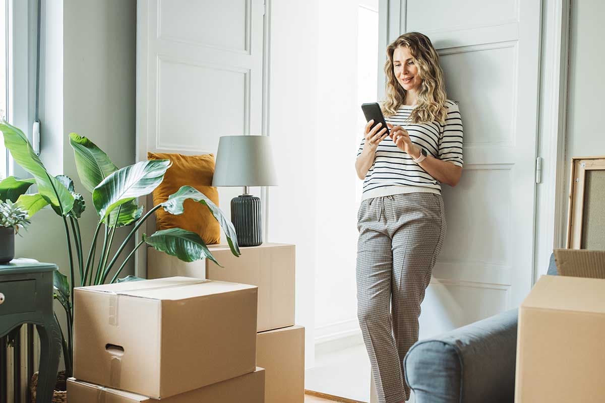 En kvinne står i et rotete rom fylt med esker, holder en telefon og ser tankefullt på omgivelsene sine