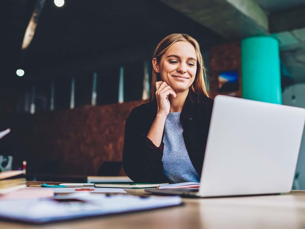 En kvinne sitter ved et skrivebord, fokusert på laptopen sin, med en notatblokk og en penn ved siden av