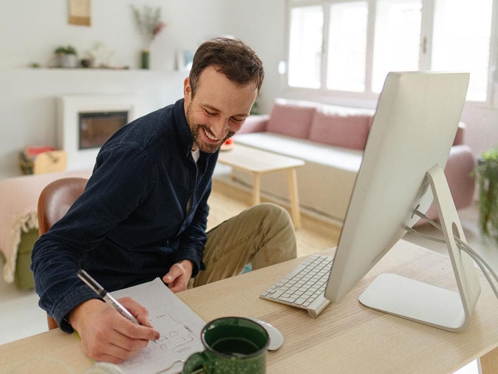 En mann sitter ved et skrivebord, fokusert på laptopen sin, med en penn i hånden, klar til å ta notater eller skrive