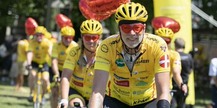 En gruppe syklister i gule skjorter og hjelmer som sykler sammen på en solrik dag